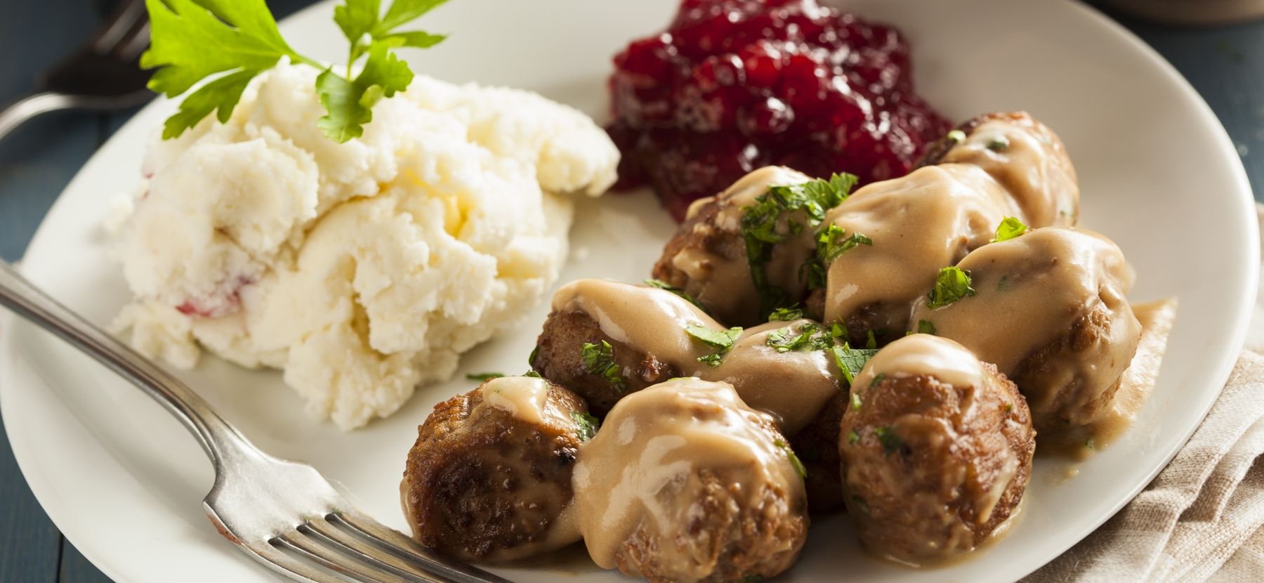 Photo of a dish of Swedish meatballs, covered with brown cream sauce, with a side of mashed potatoes and jam