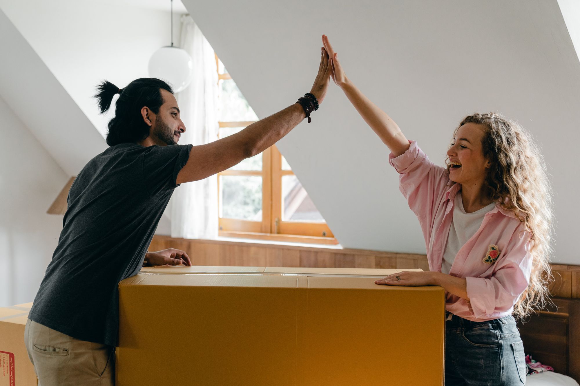 Couple are high fiving on top of an unpacked box in their new apartment