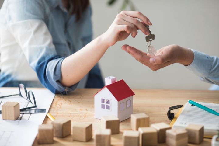 Image of a landlord handing a house key to a tenant, with a house model and paperwork shown on the desk in front of them