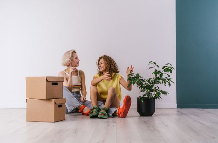 Two women sitting on the floor, next to a few boxes and a plant