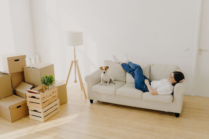 Female renter lies on white comfortable couch with a dog, next to a pile of moving boxes