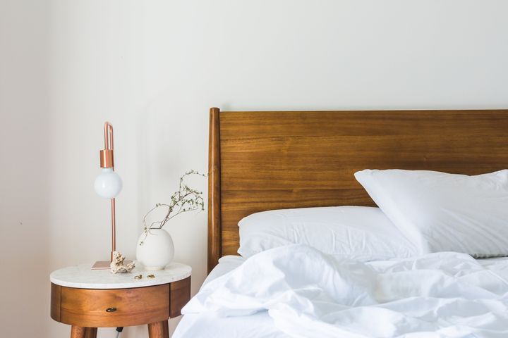 Photo of a bedroom, showing a bed with white spreadsheets, and a bedside table with a lamp and a small plant