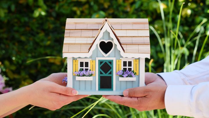 Two people holding a small model house with green plant background