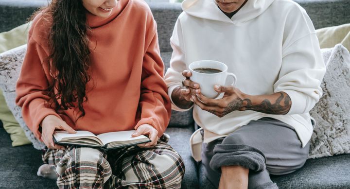 Photo of two individuals, sitting on a couch. One of them is holding a book while the other holds a coffee.