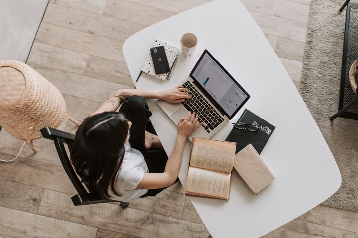 An aerial view of a woman sitting at a desk with a laptop, book, and notes