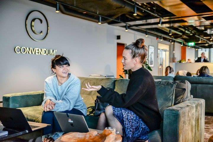 Two women sitting on a couch with their laptops open in front of them, seemingly discussing work