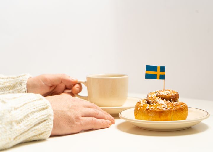 A woman holds a cup of coffee next to a pastry with a small Swedish flag on top of it