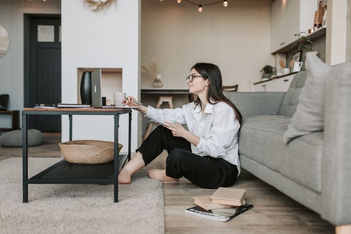 Photo of a woman sitting on the floor of her apartment, seemingly working from home, surrounded by furniture