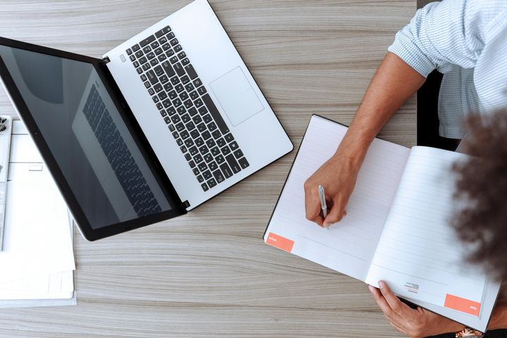 Person writing in a notepad on a desk in front of a laptop