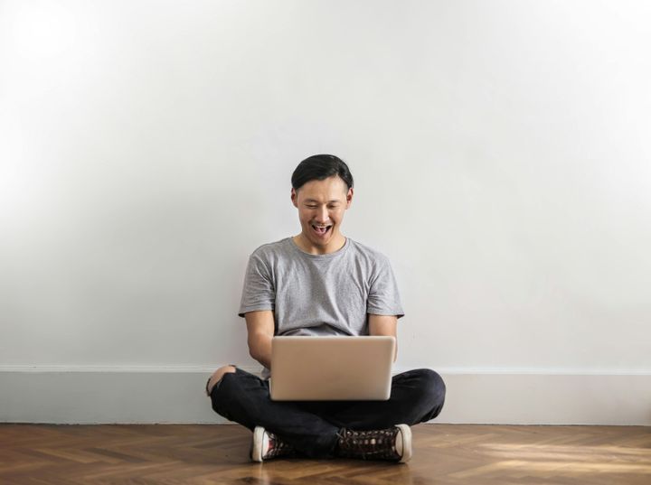 Laughing man in gray t-shirt and black jeans on sitting on wooden floor while using a laptop